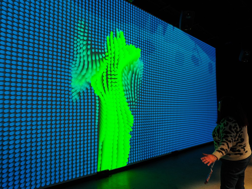A person interacts with a large digital screen displaying vibrant green and blue patterns, resembling fluid motion or waves.