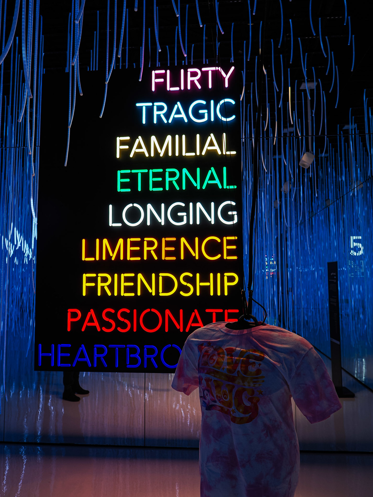 A colorful neon sign displaying various emotional terms like 'Flirty,' 'Tragic,' and 'Heartbroken,' with a pink tie-dye t-shirt hanging in front.