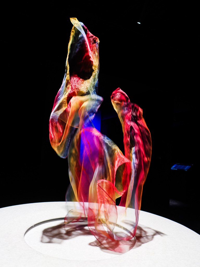 A colorful artistic sculpture made of flowing, translucent fabric, displayed against a dark background.