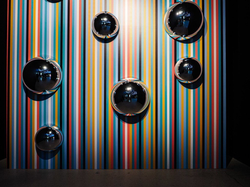 A colorful striped wall with multiple reflective circular objects mounted on it.