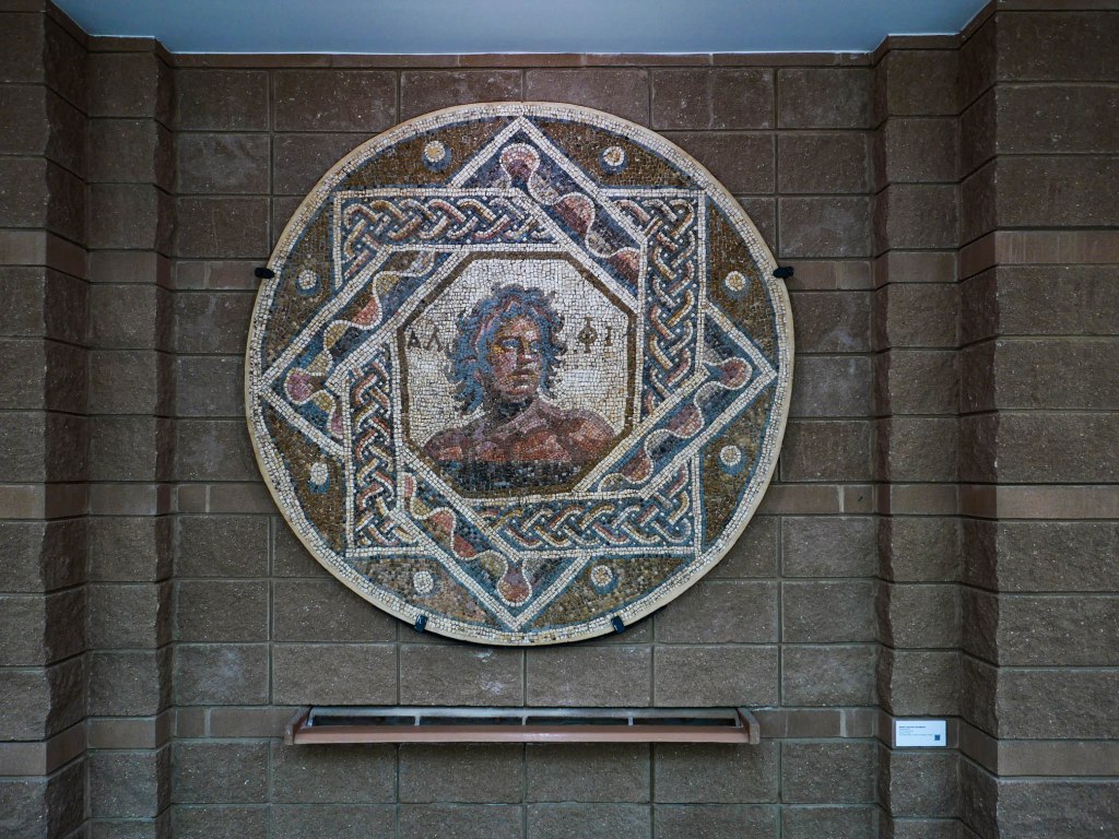 A circular mosaic featuring a portrait of a figure with curly hair, surrounded by intricate geometric patterns, displayed on a wall.