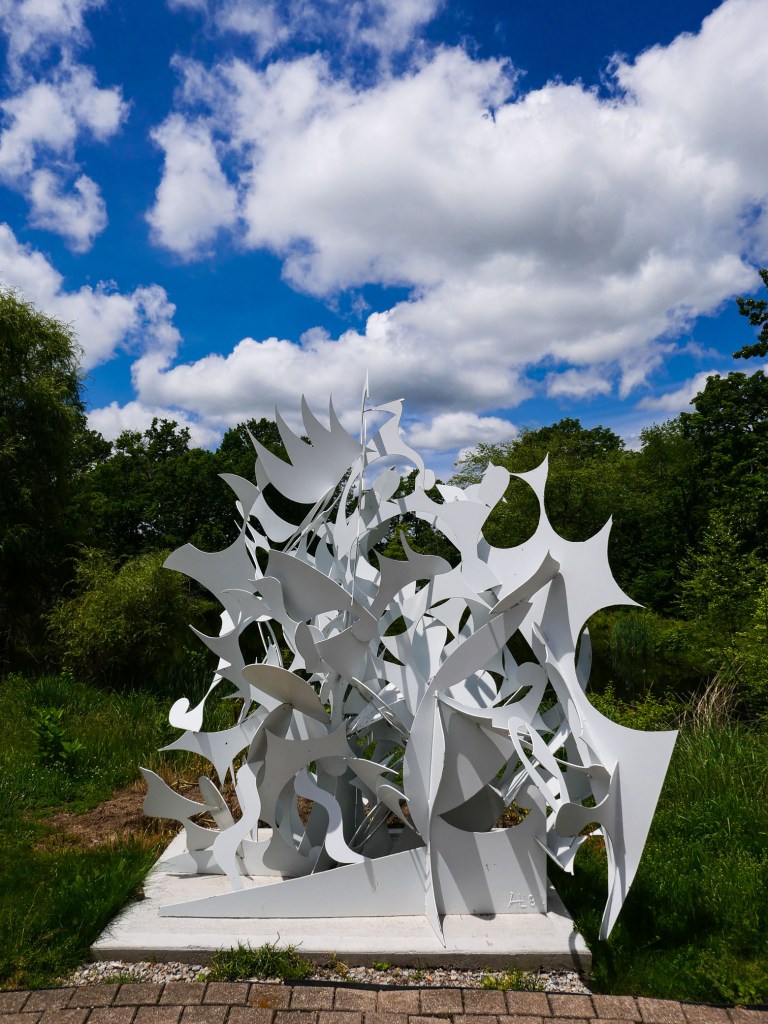 A white abstract sculpture featuring dynamic shapes and forms surrounded by lush greenery and a blue sky with clouds.