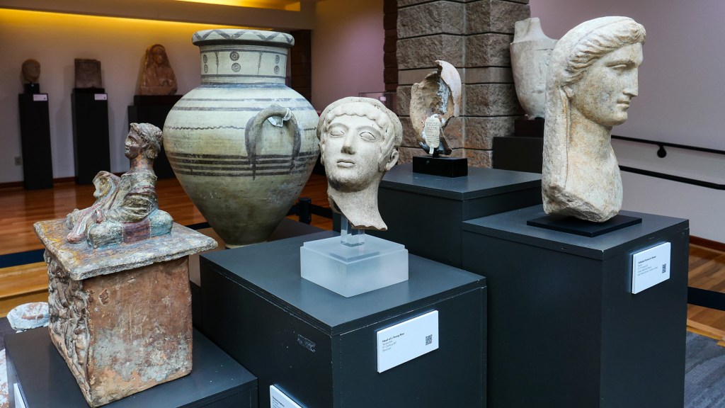 A collection of ancient artifacts displayed in a museum setting, including a large ceramic vase, sculptures of heads and figurines, and detailed pottery on black pedestals.