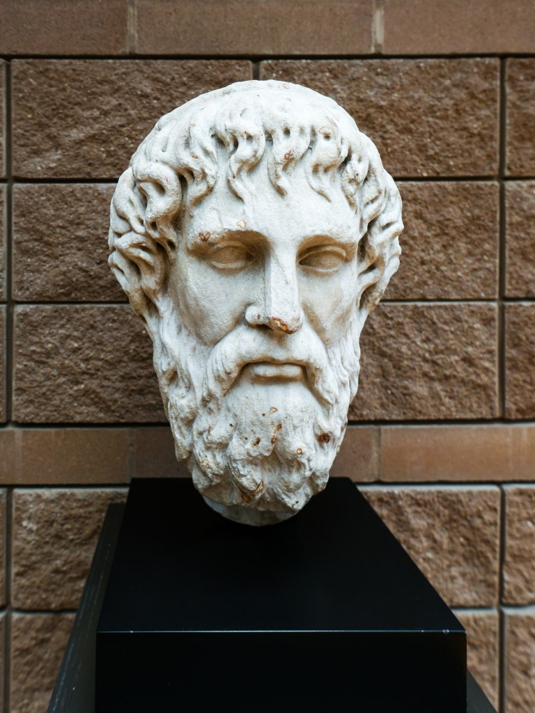A sculpted marble bust of a philosopher with long curly hair and a beard, displayed on a black pedestal against a textured stone wall.