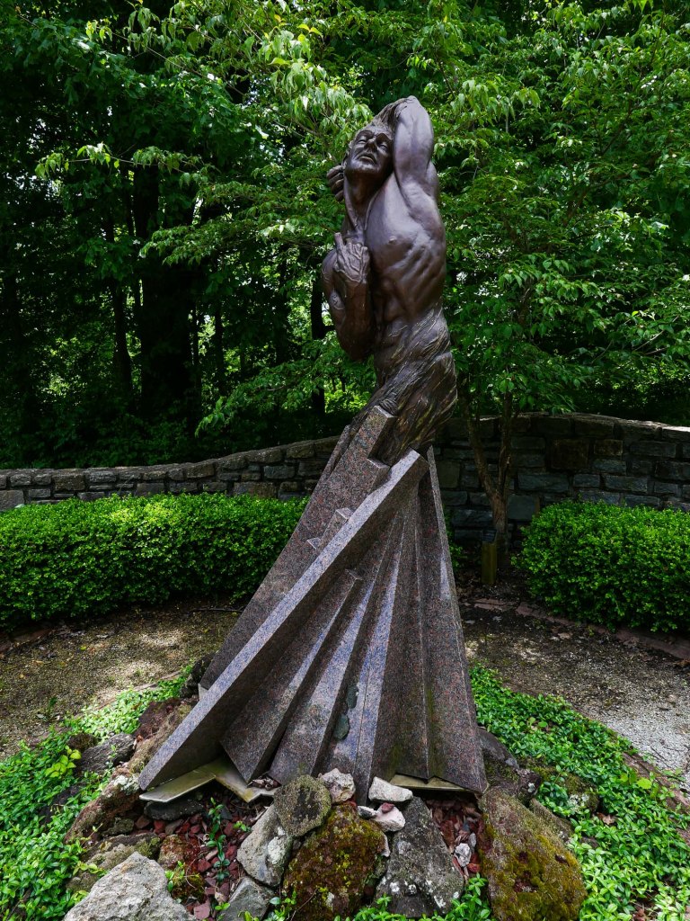 A stylized sculpture of a muscular figure in an expressive pose, surrounded by greenery and stone features.