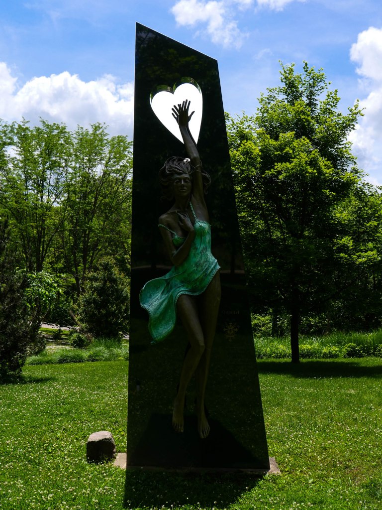 A sculpture of a woman dancing gracefully with her arm raised, set against a green landscape under a blue sky with clouds. The sculpture features a heart cutout at the top.