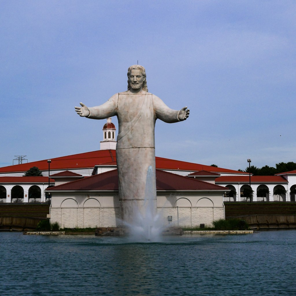 A large statue of Christ the Redeemer with outstretched arms, overlooking a body of water, with a red-roofed building in the background.