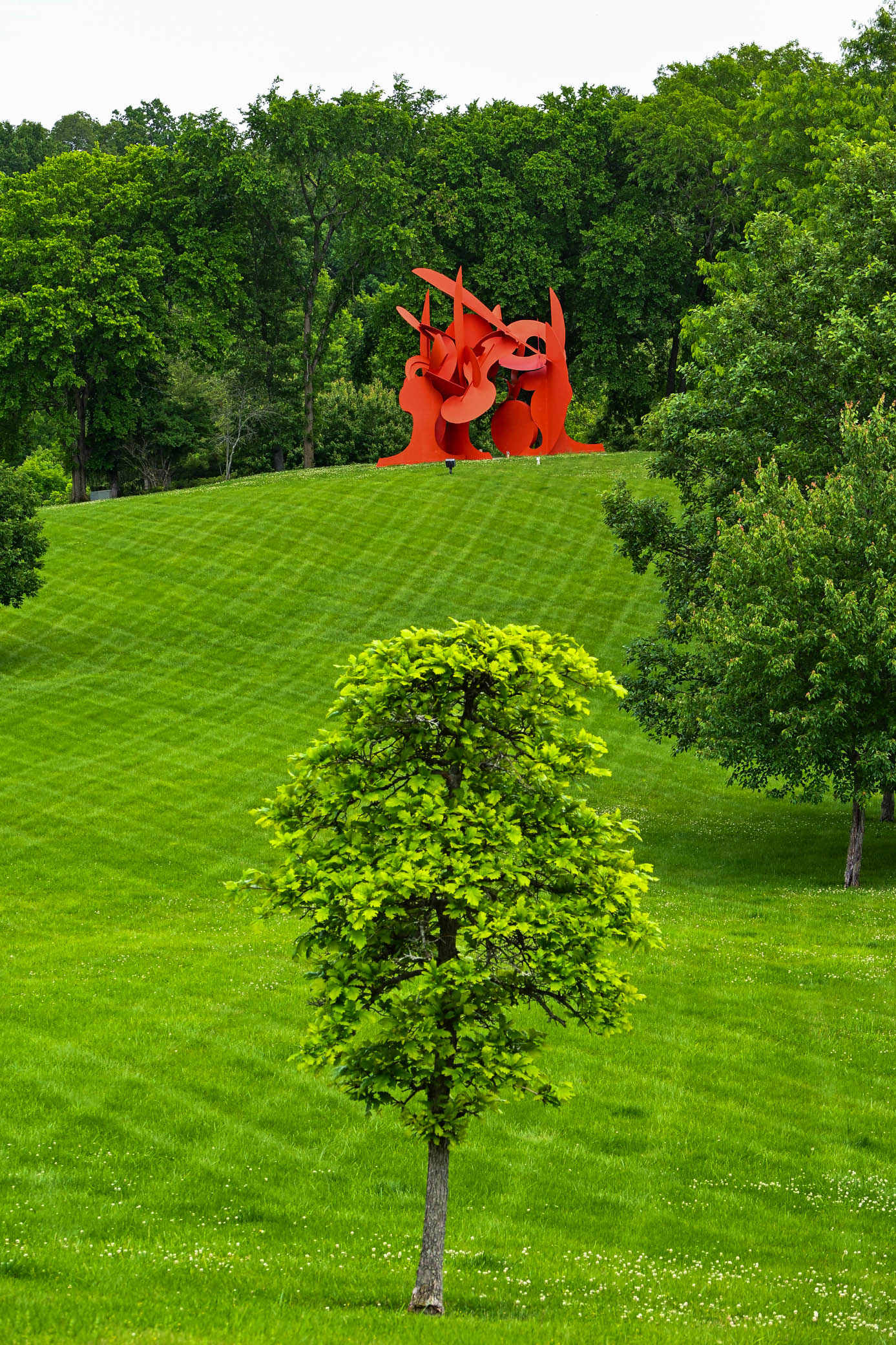 A vibrant red sculpture stands atop a grassy hill, surrounded by lush green trees.