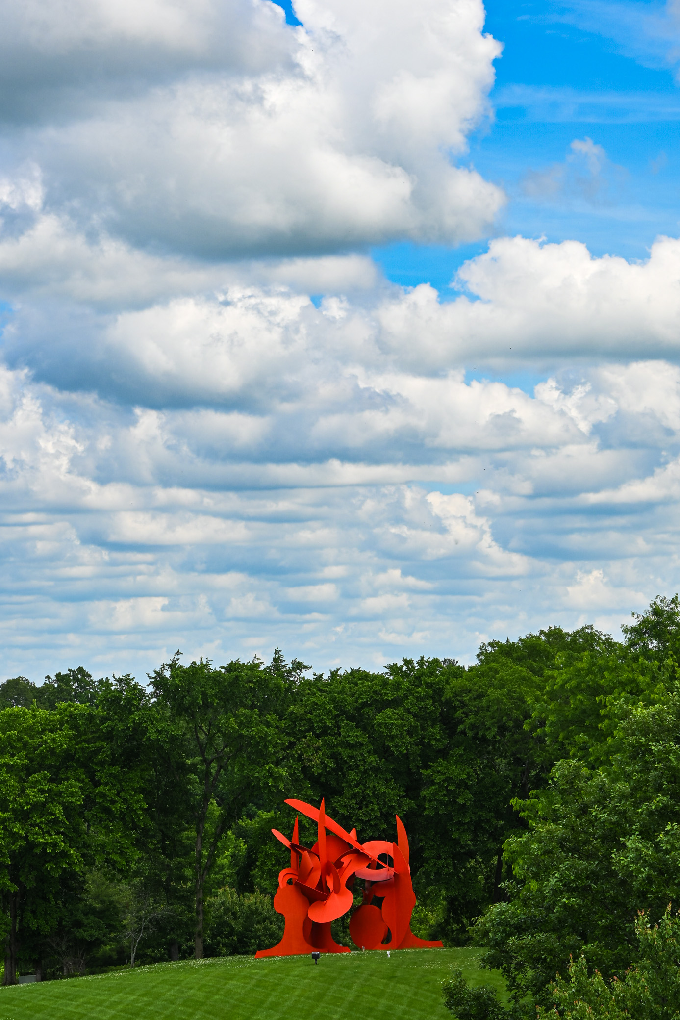 A vibrant orange abstract sculpture set against a backdrop of lush green trees and a blue sky with fluffy white clouds.