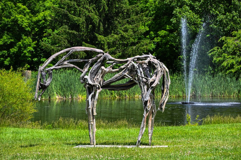 A sculpture of a horse made from twisted and intertwined branches, located near a pond with a fountain and lush greenery in the background.