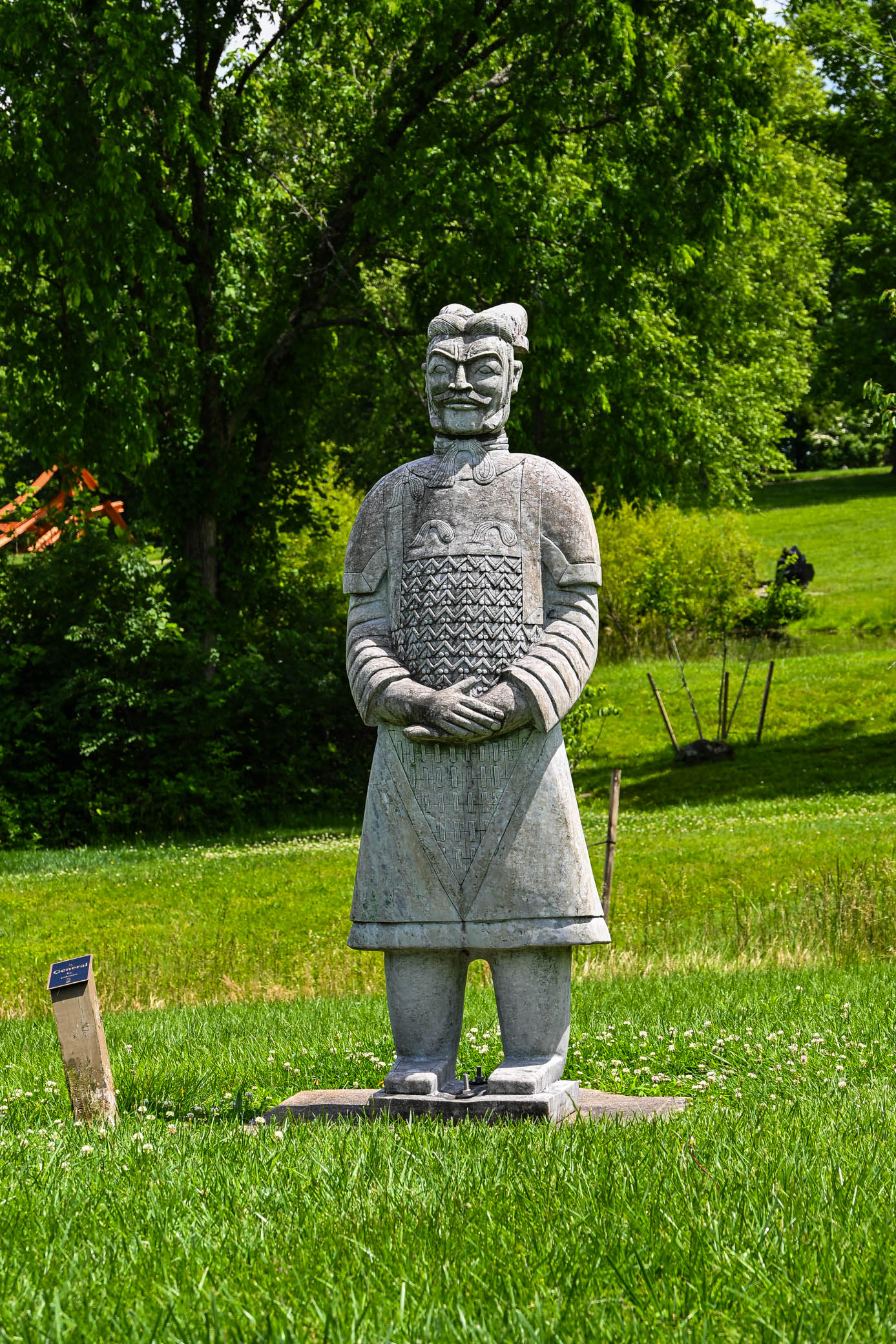 A stone statue of a warrior dressed in armor, with a fierce expression, standing on green grass surrounded by trees.