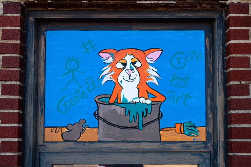 A mural featuring an orange cartoon cat playfully sitting in a paint bucket, with blue paint dripping over the sides. The background is bright blue and includes the words '#Create' and 'art', along with a simple stick figure and a small mouse illustration.
