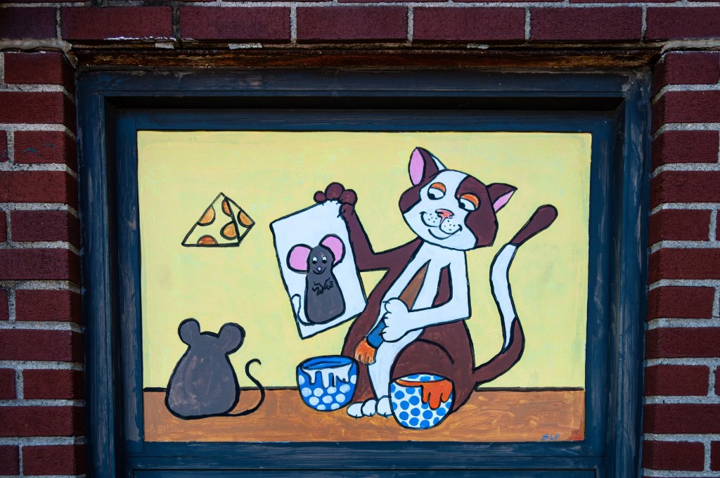 A whimsical mural featuring a cartoon cat holding a picture of a mouse, with a cheese wedge in the background. A mouse silhouette is visible in the foreground alongside two paint buckets.
