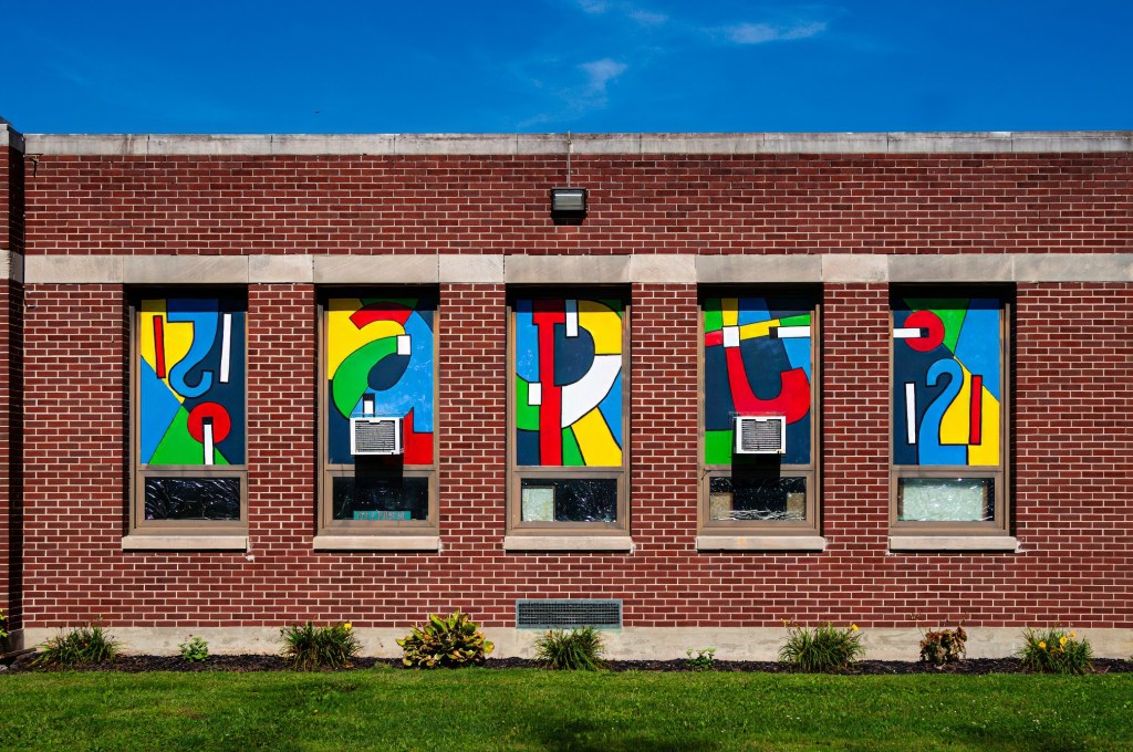 Colorful abstract stained glass windows featuring various geometric shapes and letters, set against a red brick wall.