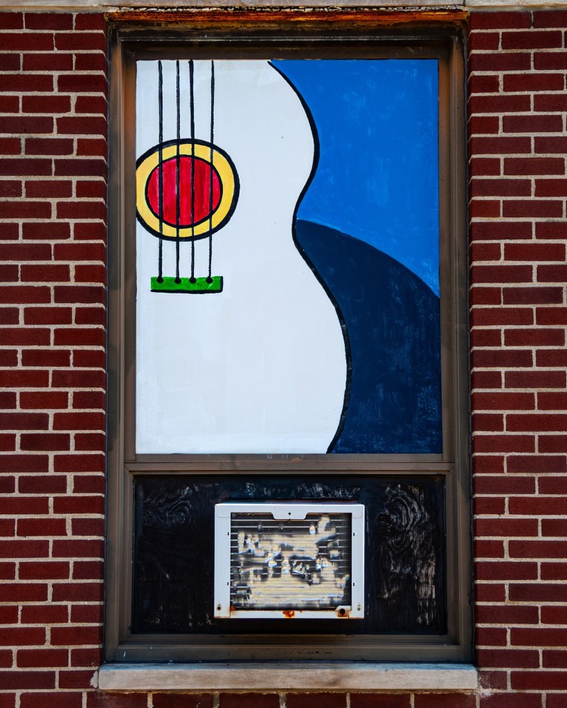A colorful mural in a window featuring an abstract guitar design with a red and yellow circular shape, set against a blue background. Below the mural, there is an air conditioning unit.